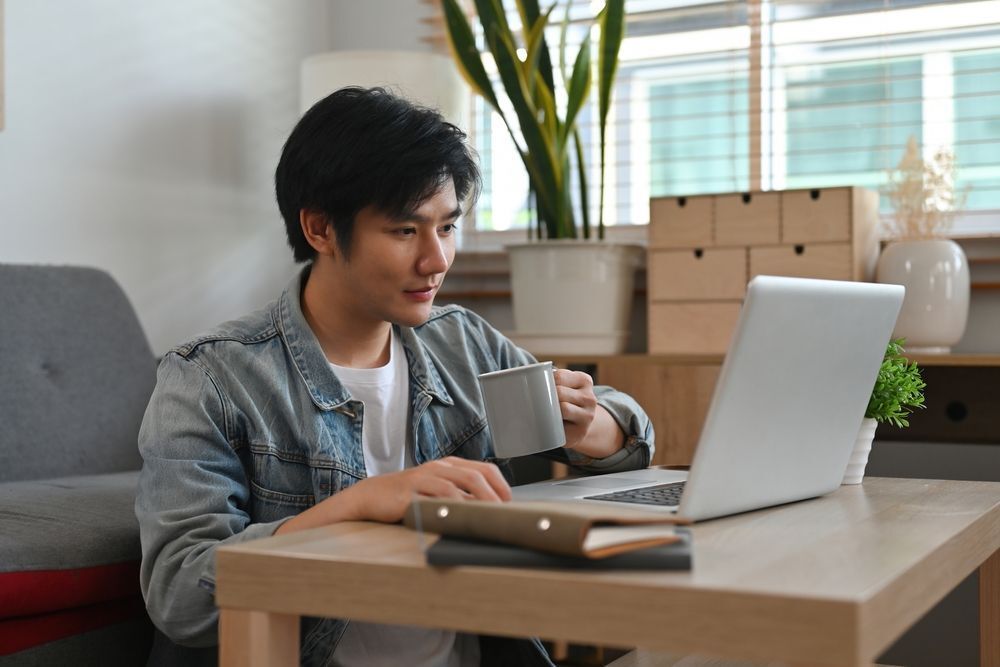 A young man holding a cup while working from home in his living room.