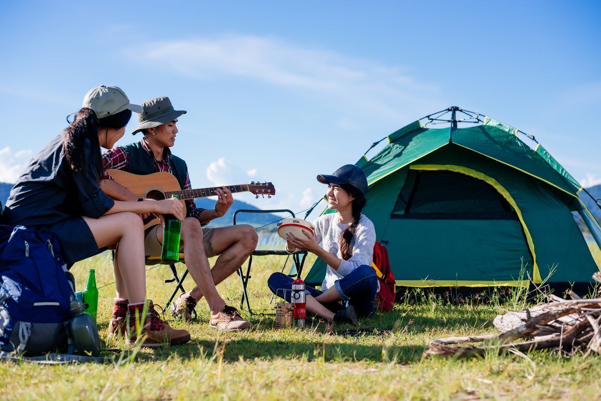 Man playing a guitar while two female friends happily listening with a visible mountain view and tent.