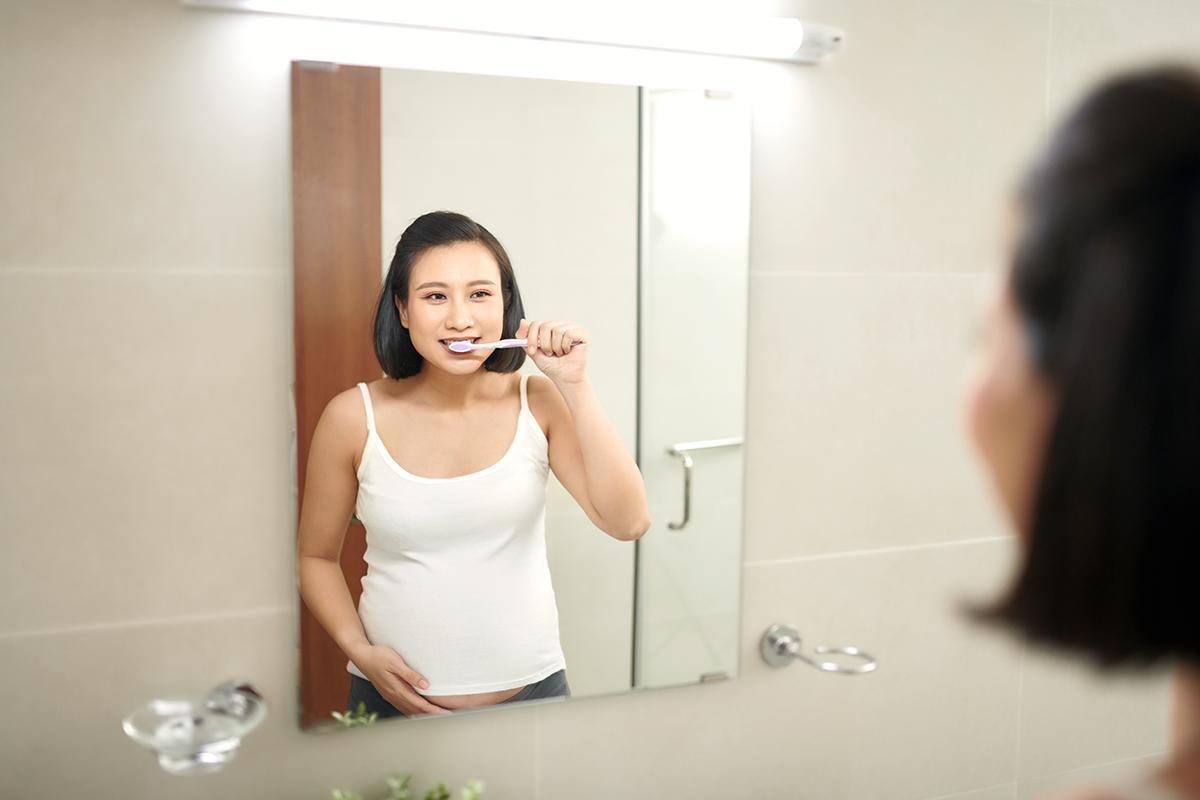 A pregnant Asian woman brushing her teeth in front of a mirror.