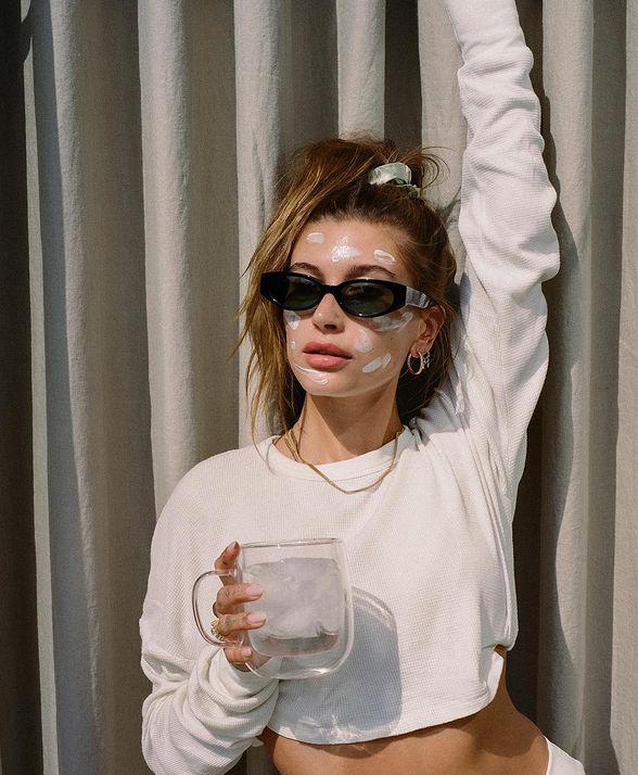 Woman holding a glass while wearing a white sweater, sunglasses, and sunscreen 