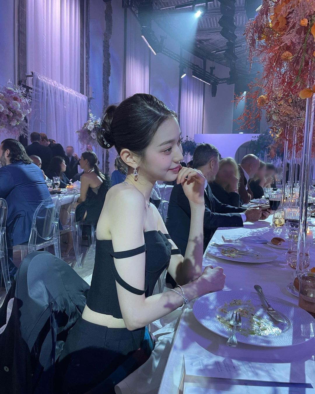 IVE's Wonyoung in a black dress sitting in front of a white dinner table.