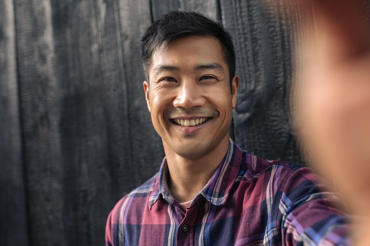 Young Asian man in plaid taking a selfie against a dark wooden background. 