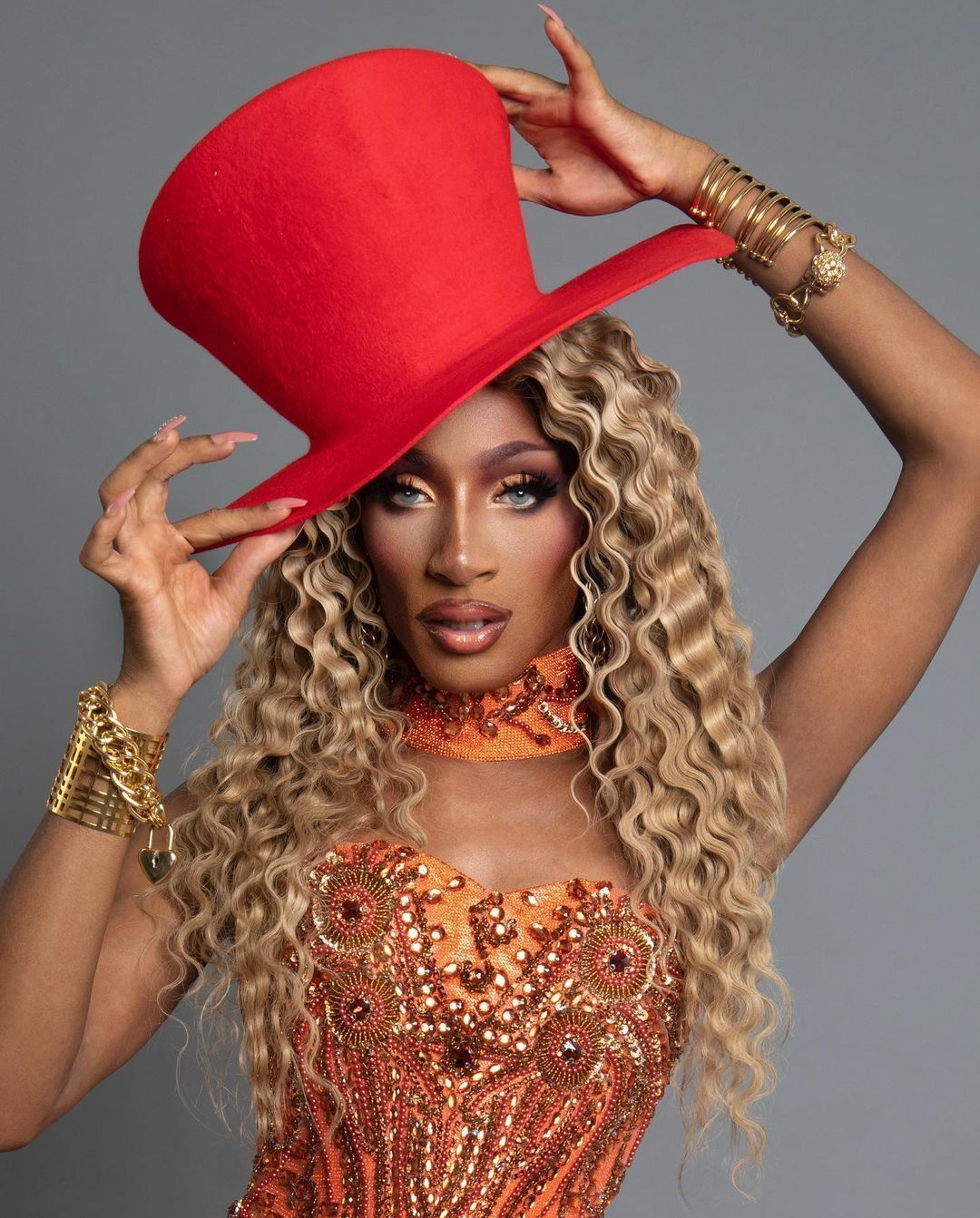 A portrait of drag queen with blonde curly wig and big, red hat.