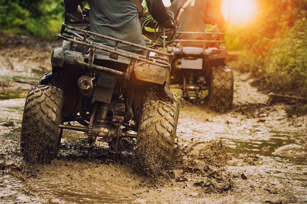 Two men driving ATVs on a muddy trail outdoors.