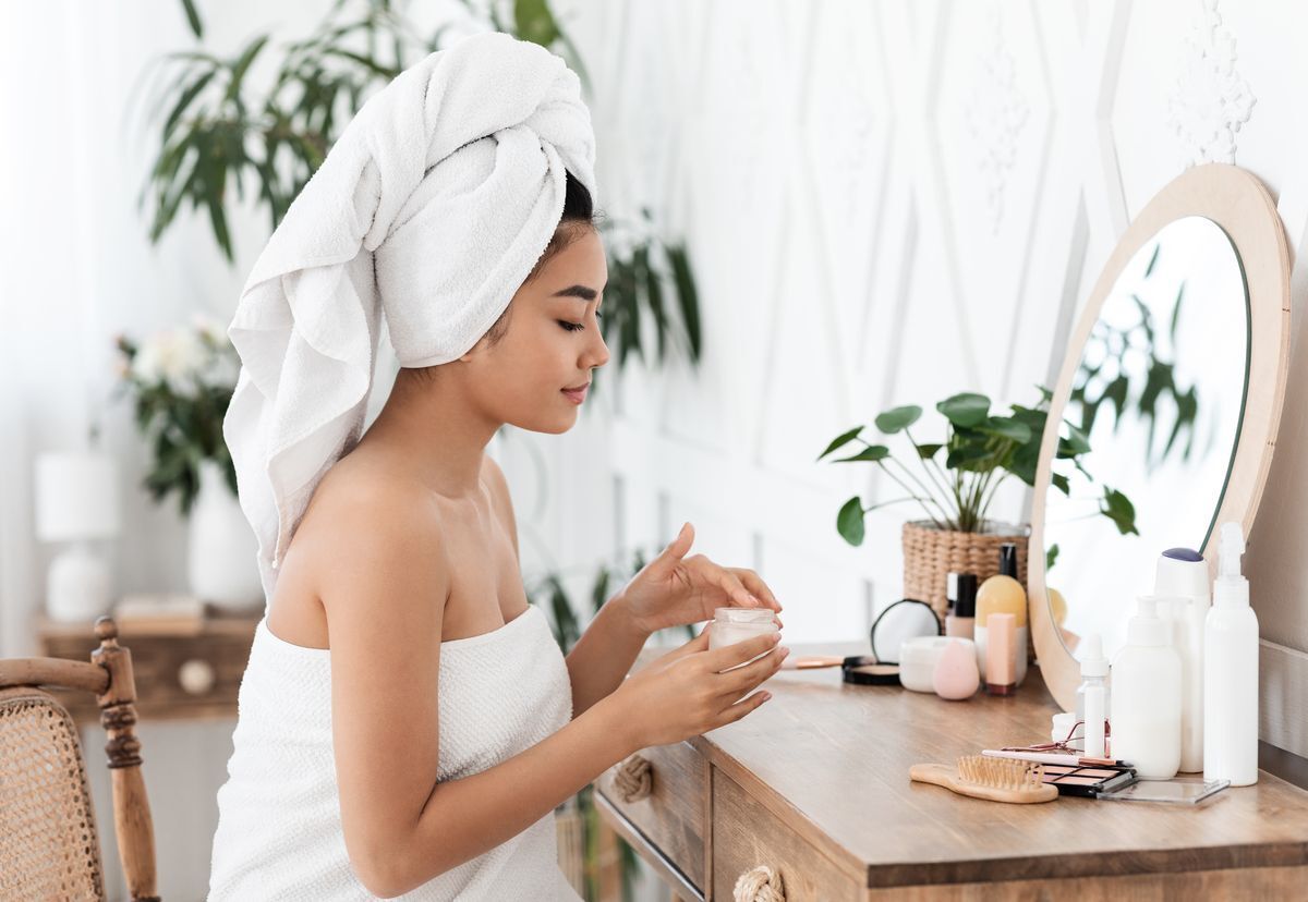 Asian woman in a towel applying moisturizer in front of a mirror.