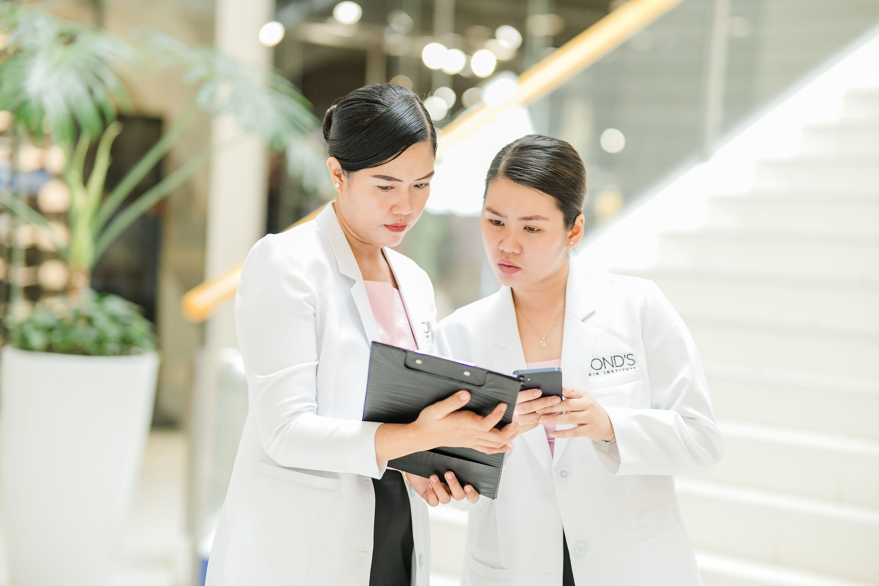 Two women in lab coat talking while looking at clipboard.