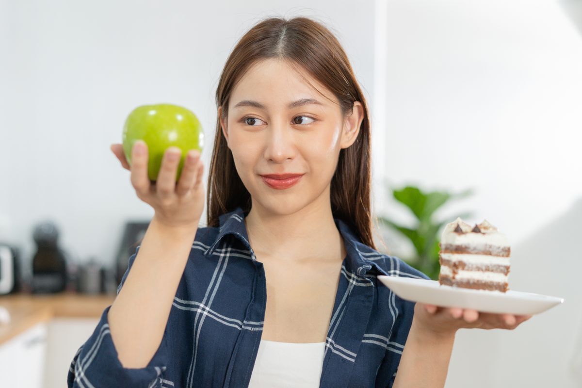Asian girl smiling and looking at the apple she’s holding with one hand, with a plate of cake in the other hand.