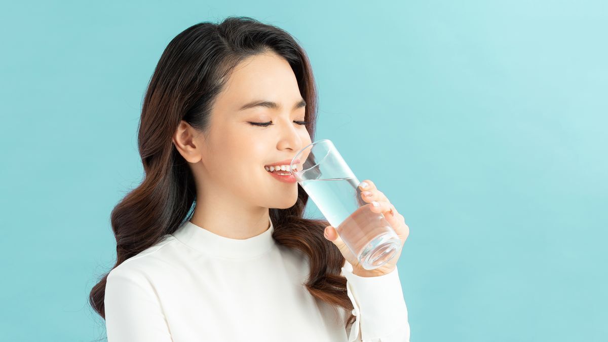 Asian girl smiling and drinking a glass of water against a blue background