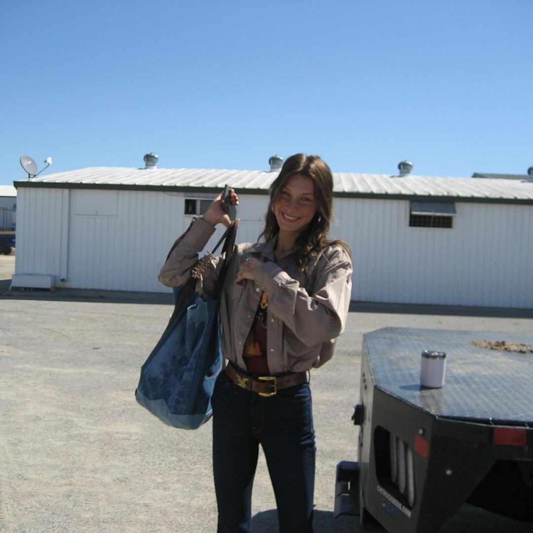 Bella Hadid in a bomber jacket, denim pants, and a big belt smiling at the camera.