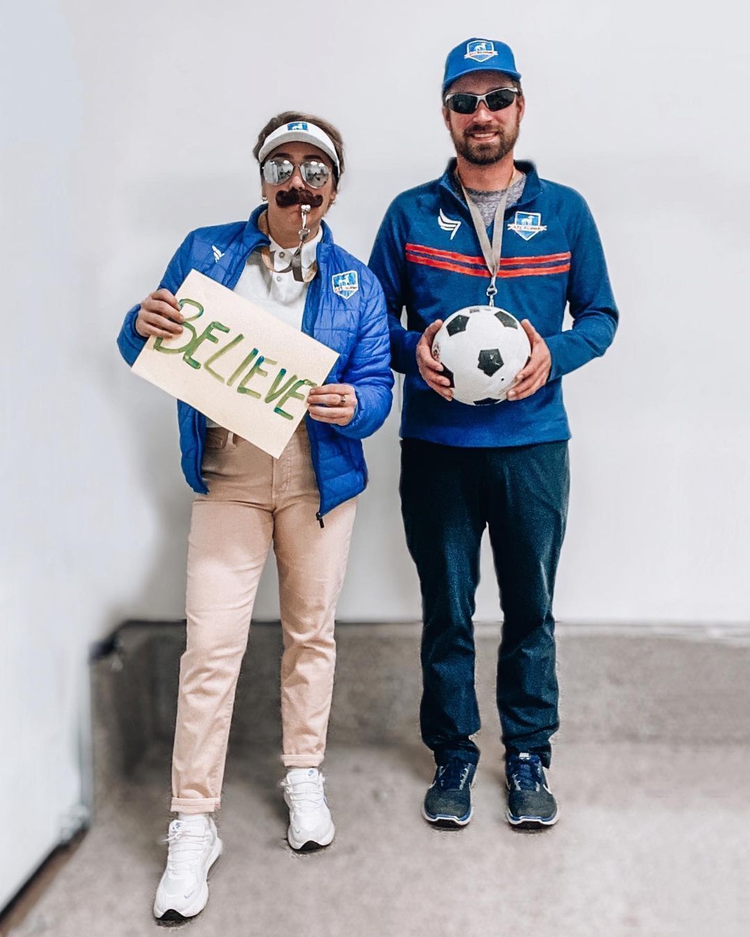 A woman dressed up as Ted Lasso blowing whistle and holding a ‘Believe’ poster.