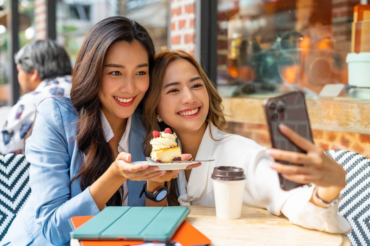 Asian woman in a white shirt taking a selfie with another woman in a blue blazer holding up a slice of cake.