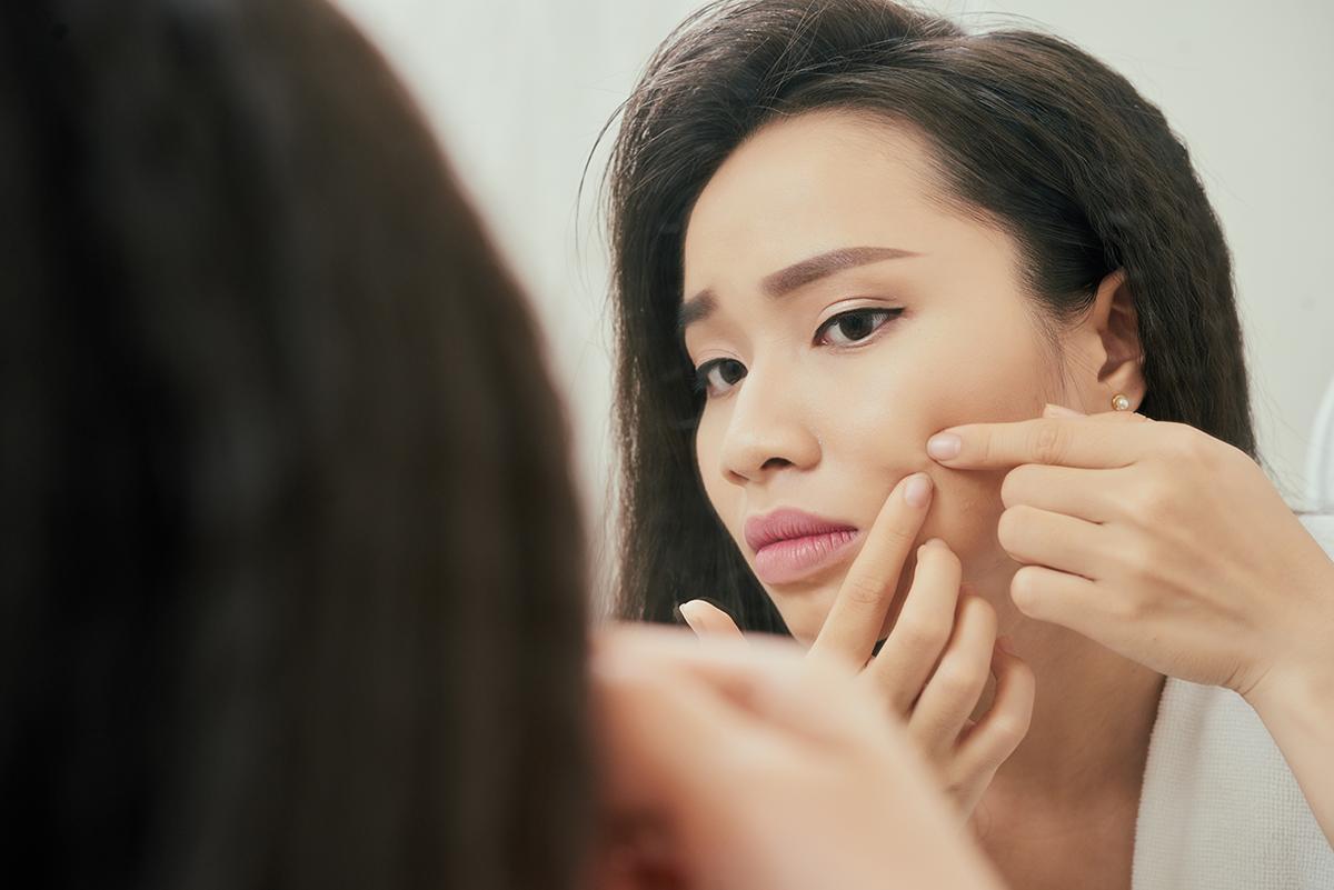 Asian woman popping pimple in front of a mirror