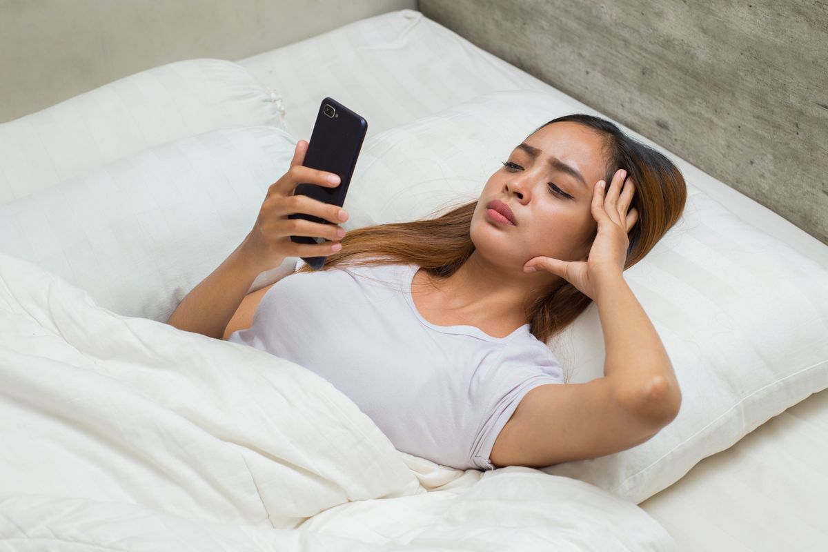 Filipino woman lying in bed looking worried as she holds her phone.