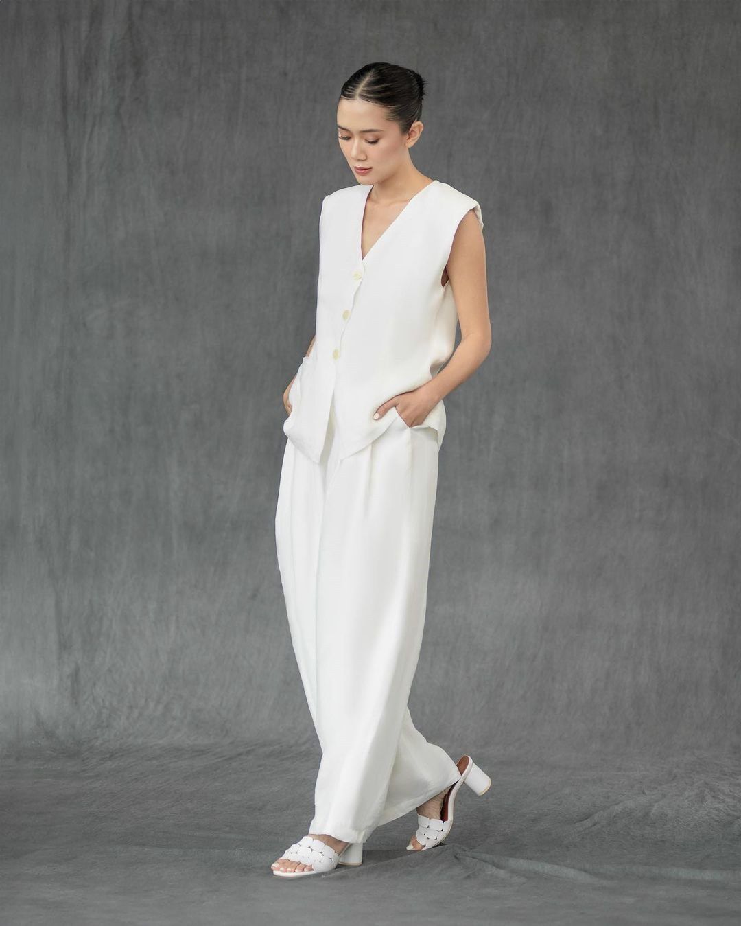 Asian woman wearing sleeveless white pantsuit against gray wall.