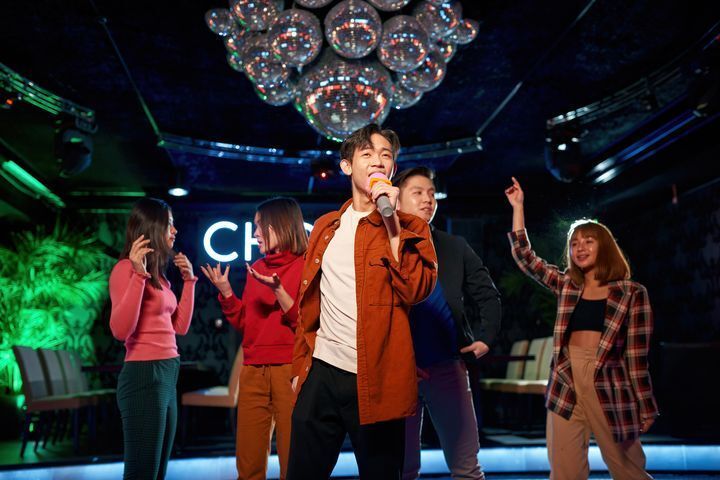 Filipino man singing karaoke at a party, with colleagues in the background.