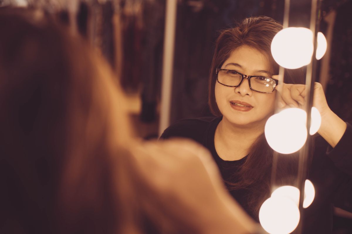 Middle aged Filipino woman with glasses looking at lit mirror.