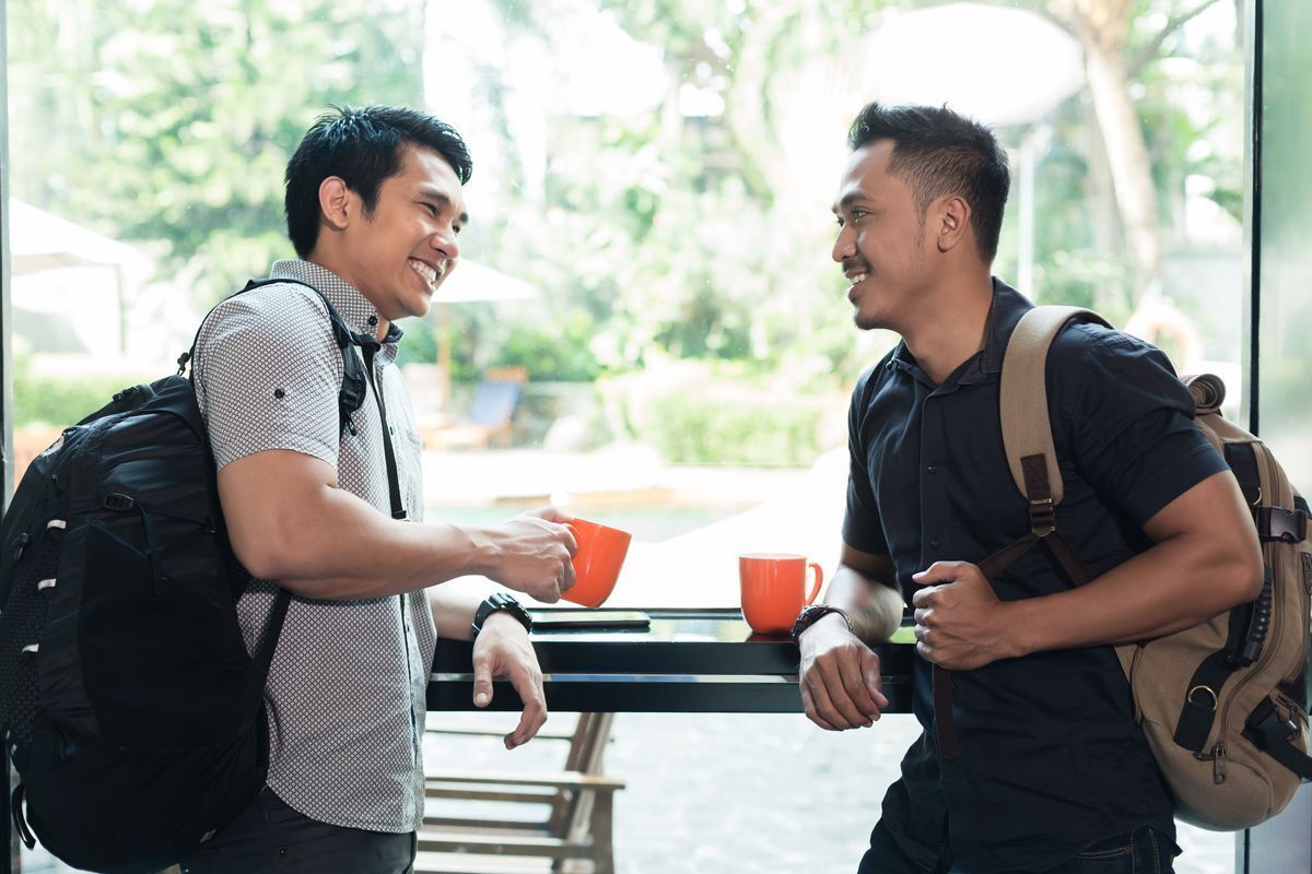 Two Asian men with backpacks happily drinking coffee together against a window.
