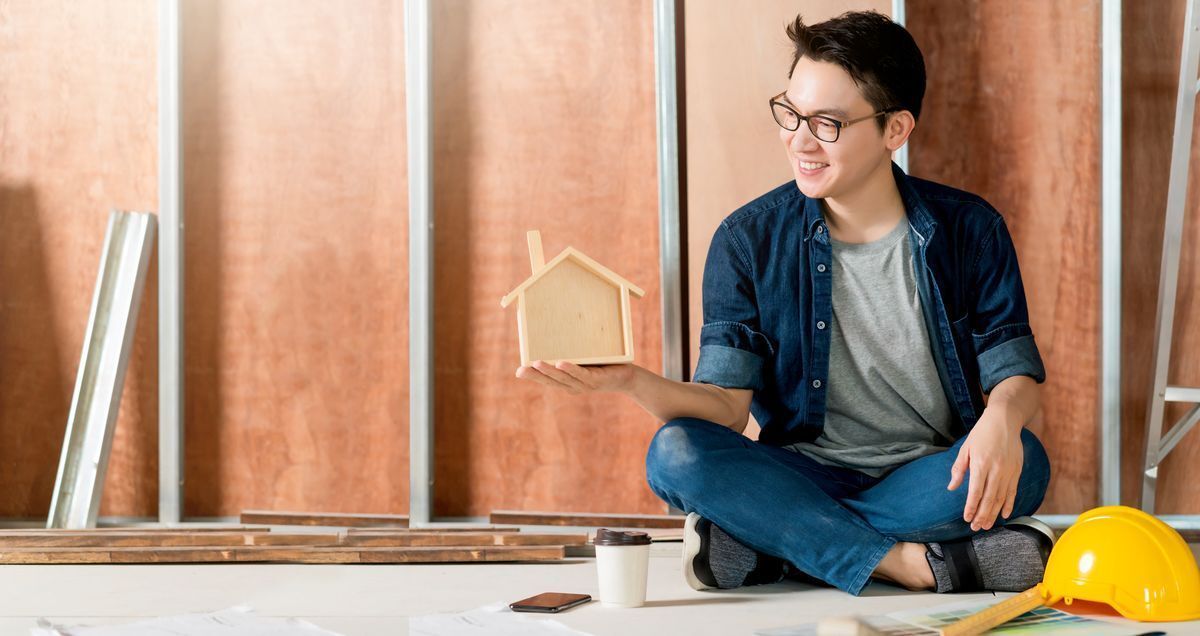 Smiling Asian man in glasses and denim holding a wooden house scale model.