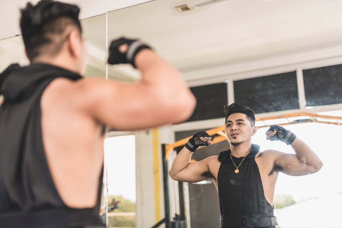 Asian man flexing his biceps at the gym while facing a mirror.