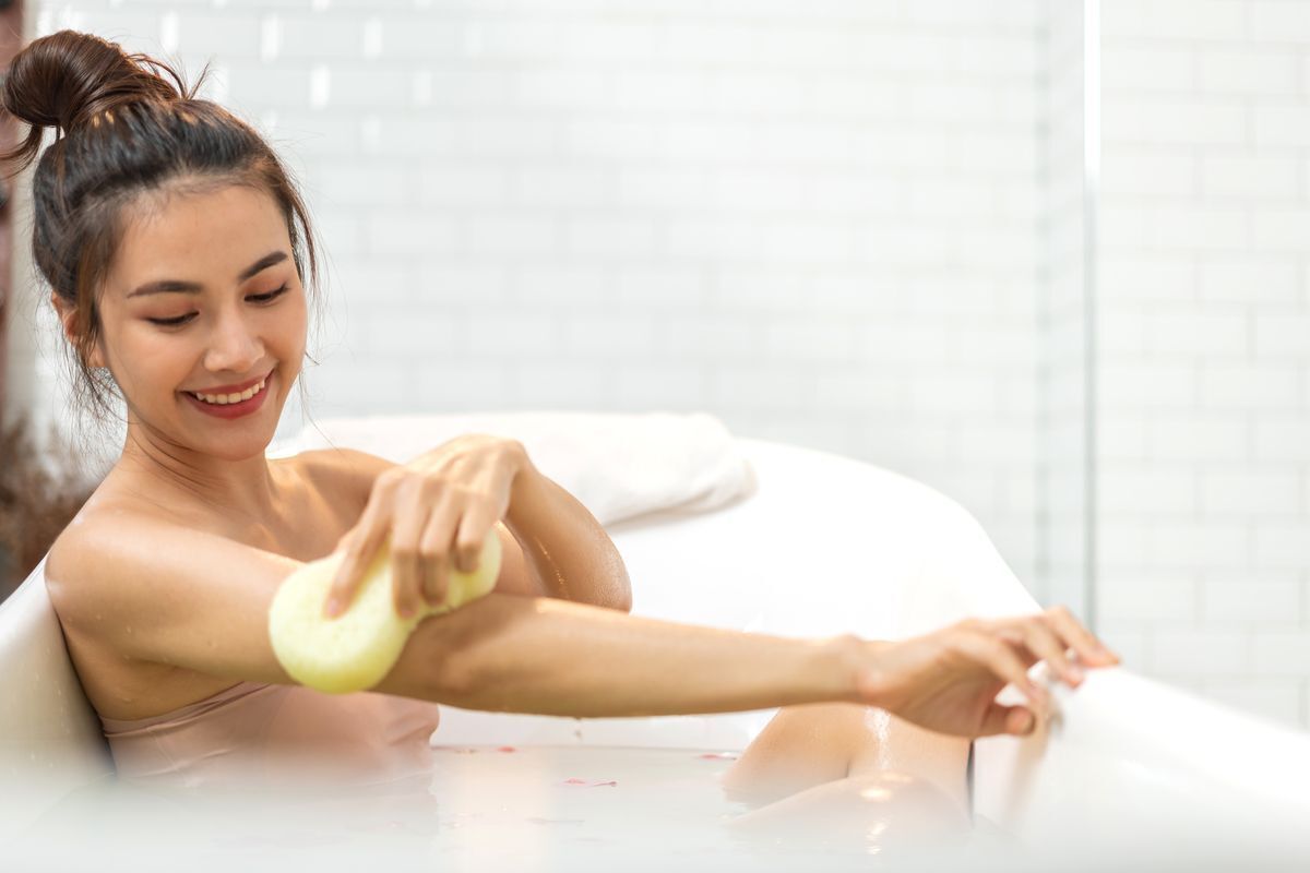 Woman in a bathtub cleaning her arm with a wet sponge.