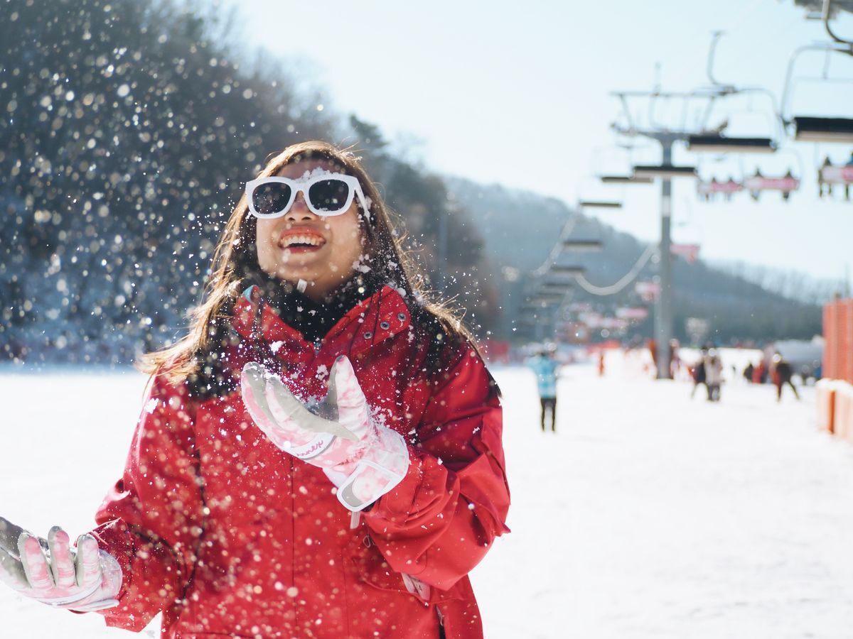 Asian woman in sunglasses and a red winter coat, smiling against cable cars and a snowy background while snow falls.