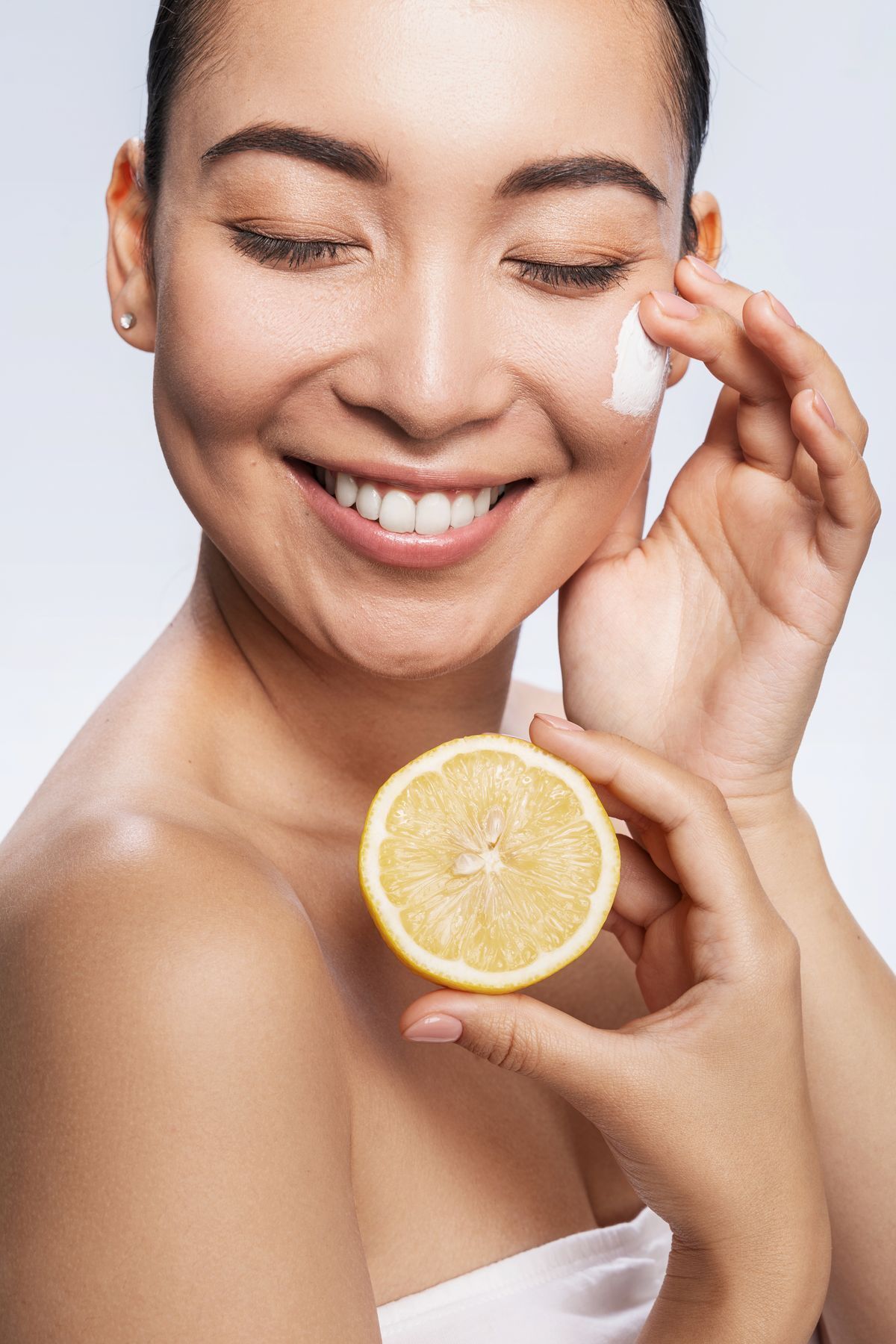 Asian woman smiling and applying cream to face while holding up a sliced lemon.