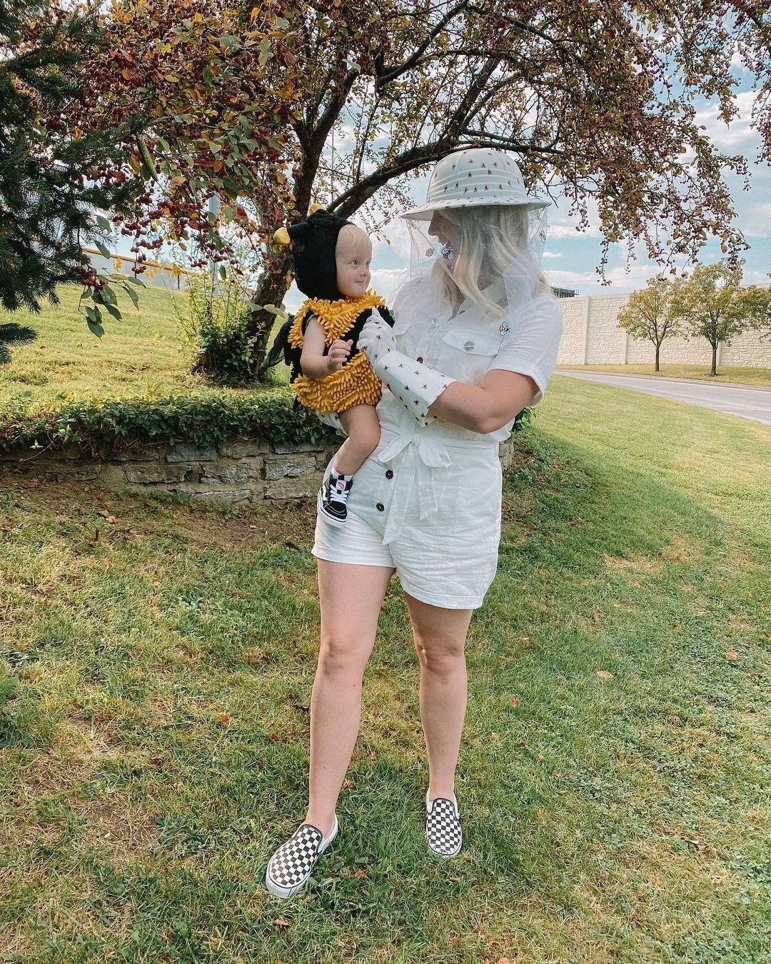 Woman wearing white romper and beekeeping veil holding a baby dressed in bee costume.