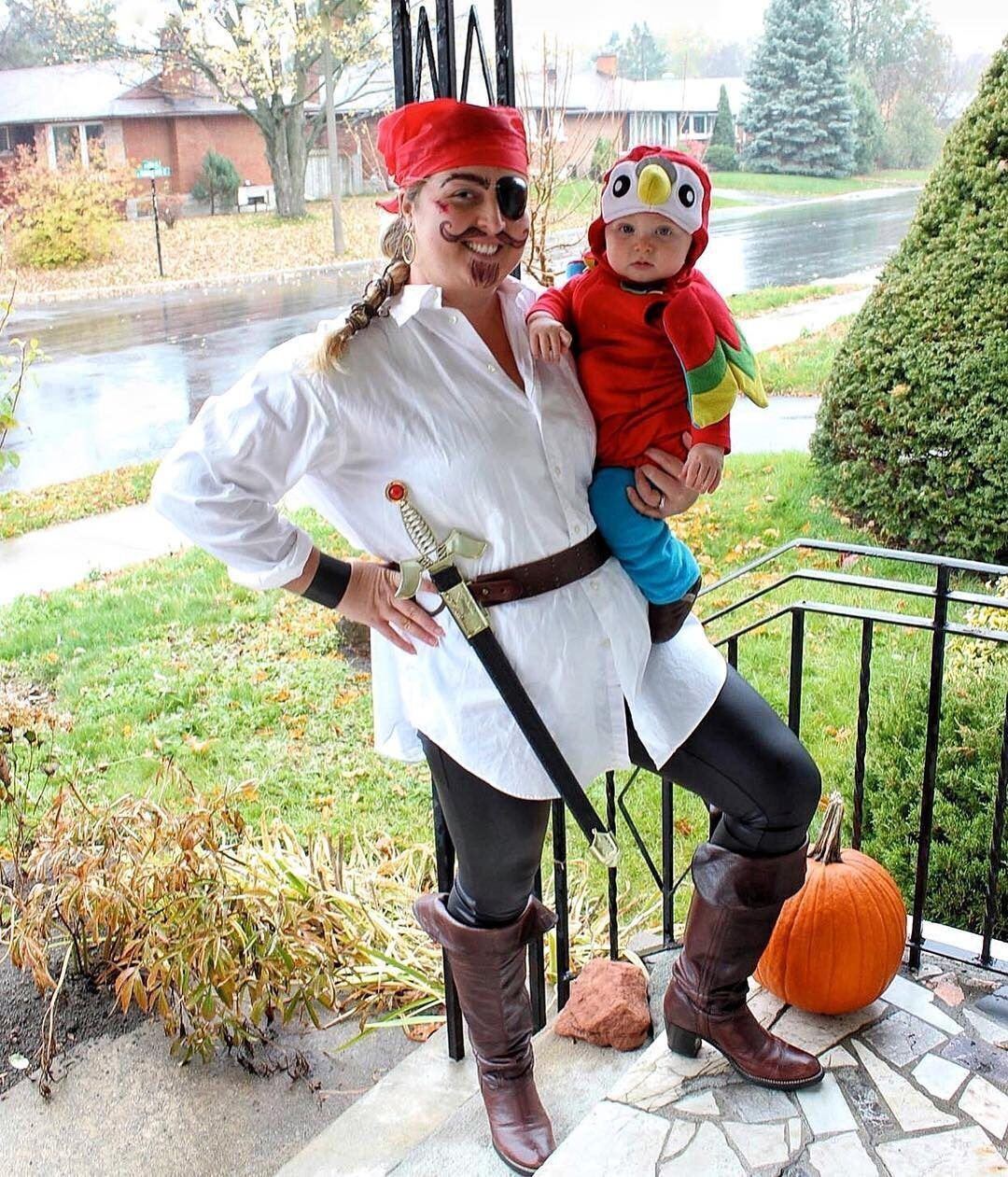 Woman posing as a pirate with an eyepatch while holding a baby dressed in a parrot costume.