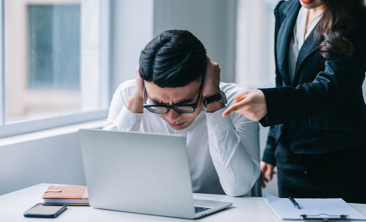 Asian man with glasses, covering ears while looking at laptop with woman pointing finger behind him.