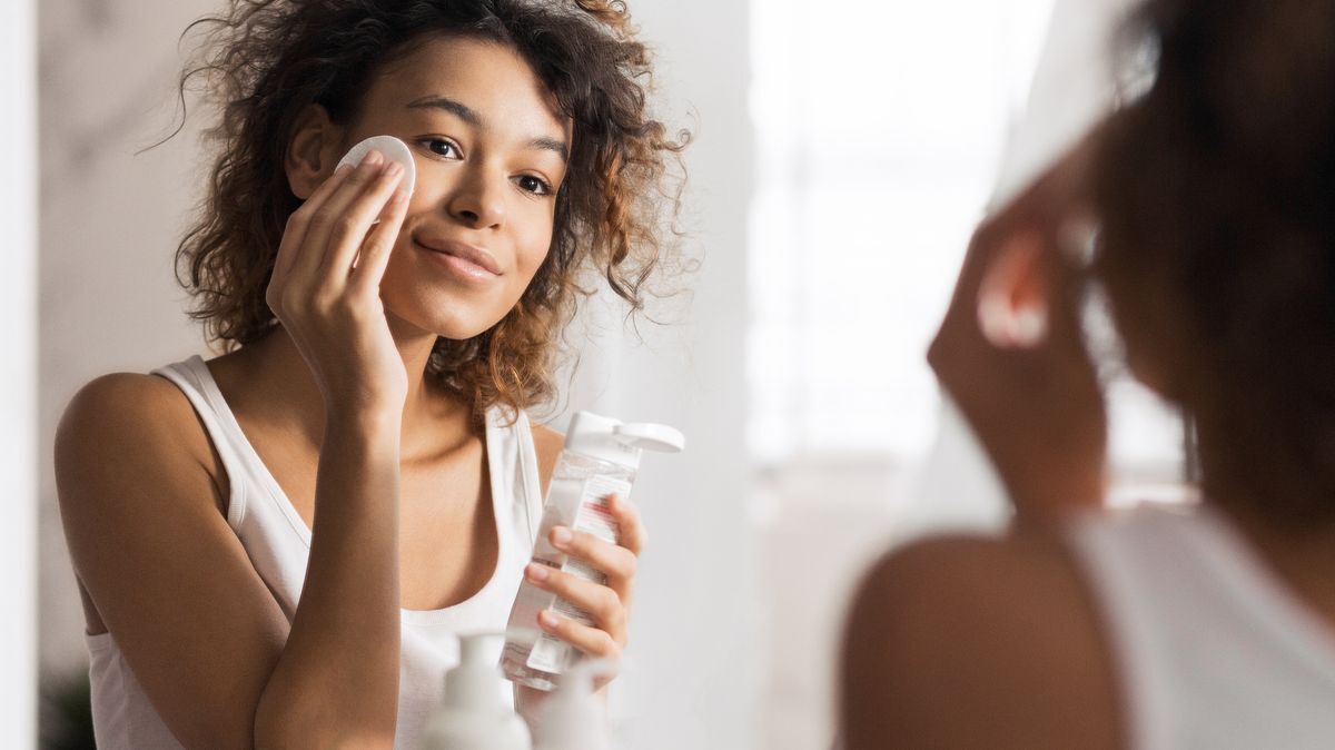 Woman with curly hair and dark skin exfoliating her skin with a cotton pad. 