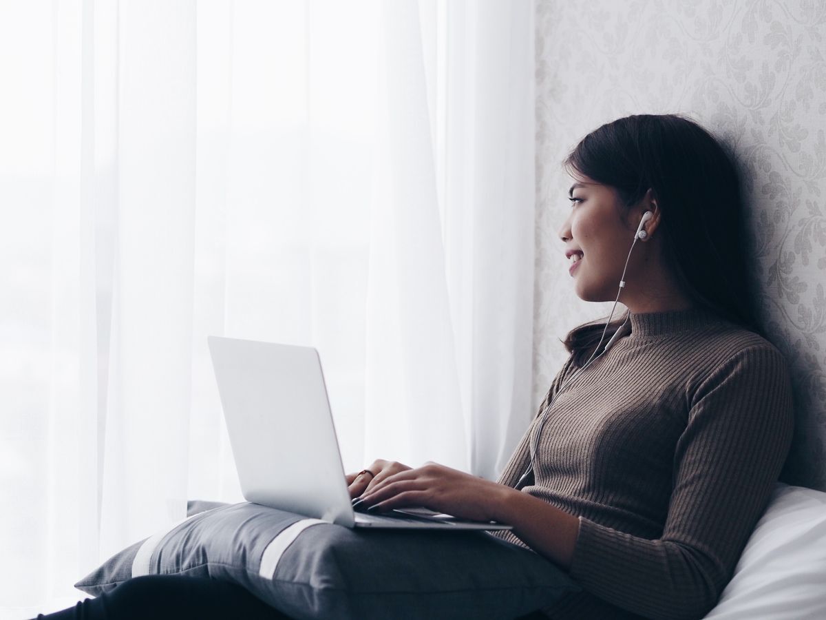 Filipino woman smiling, looking at window while on the bed with a laptop and earphones 