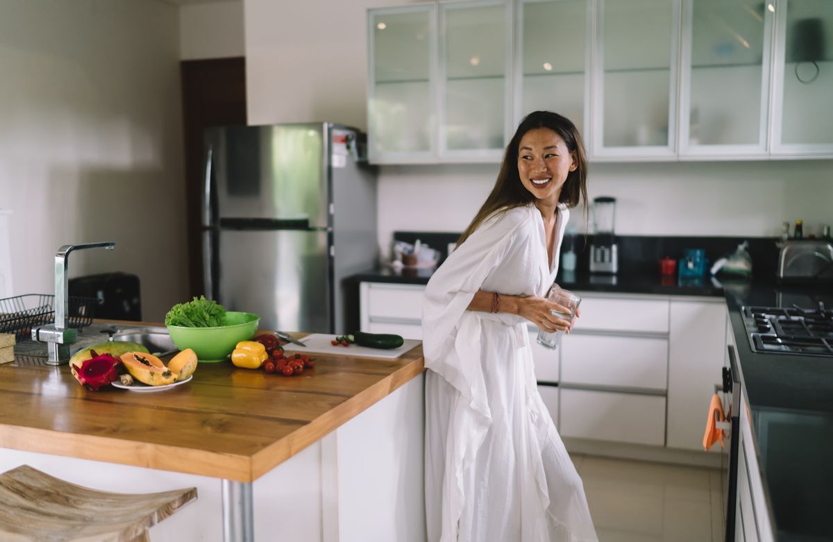 Asian woman wearing a white dress, holding a glass of water in the kitchen.