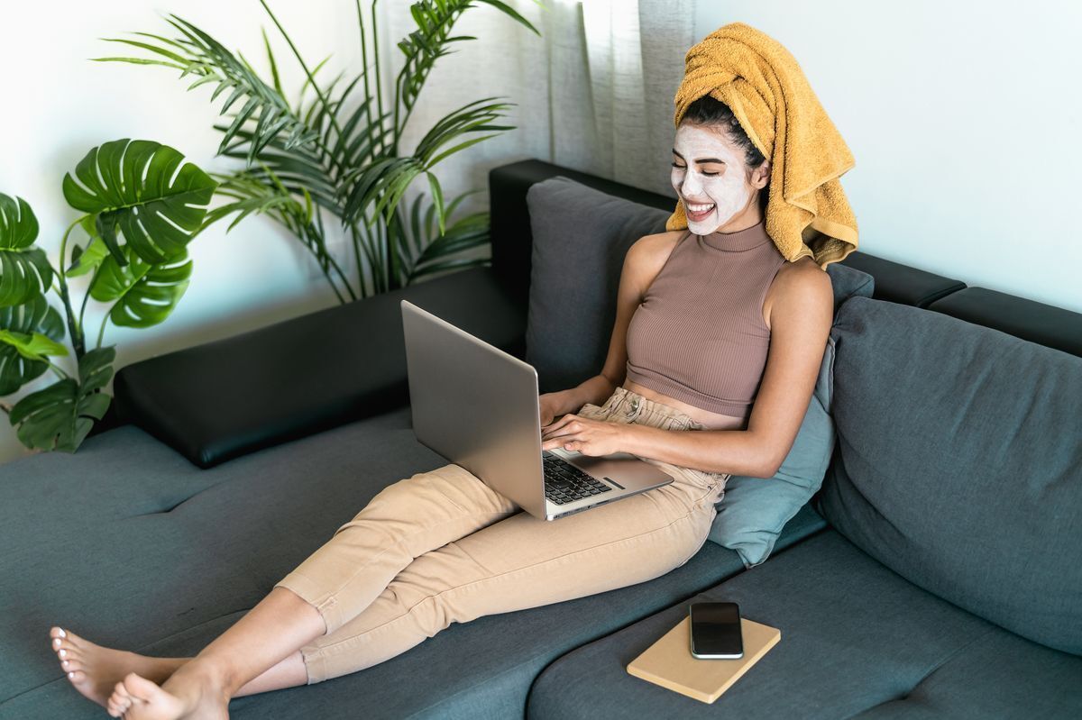 Asia woman with a face mask and towel on head sitting on a couch with her legs up, while working on a laptop.