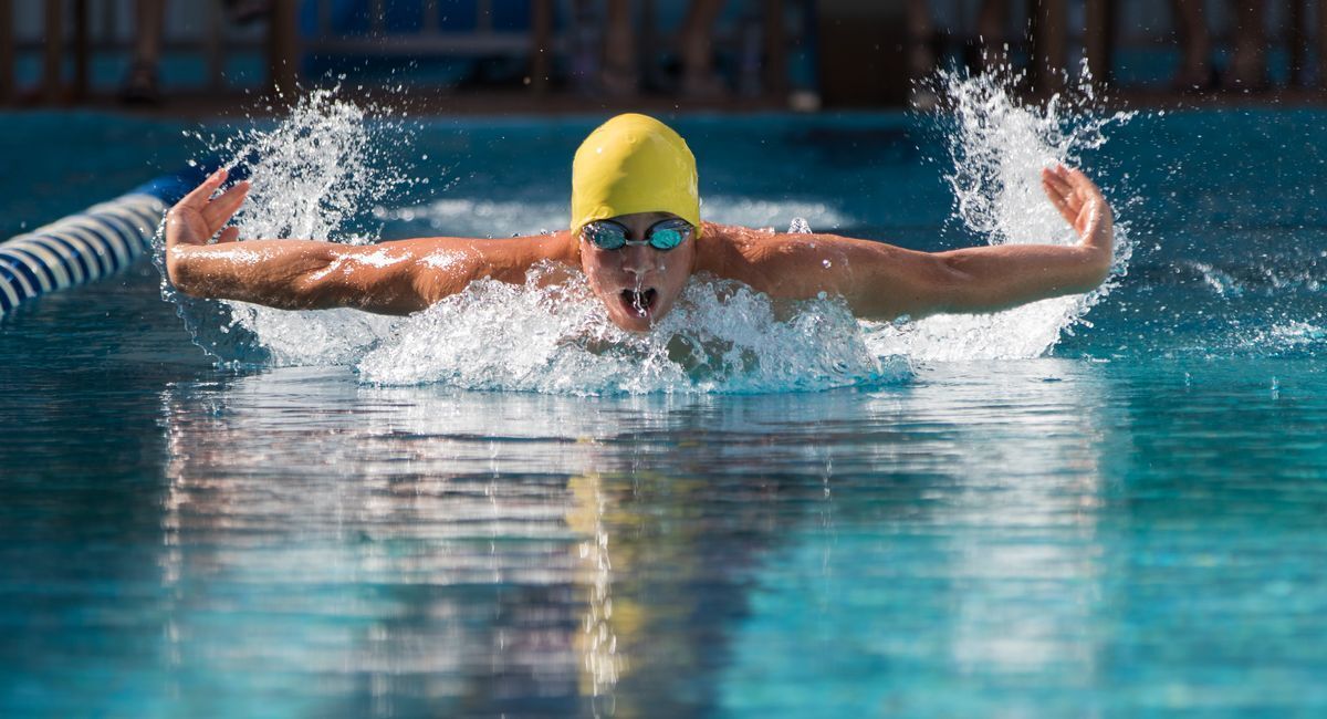 Male swimmer swimming in a lap pool wearing a yellow swim cap and goggles with visible arms and water splashing. 