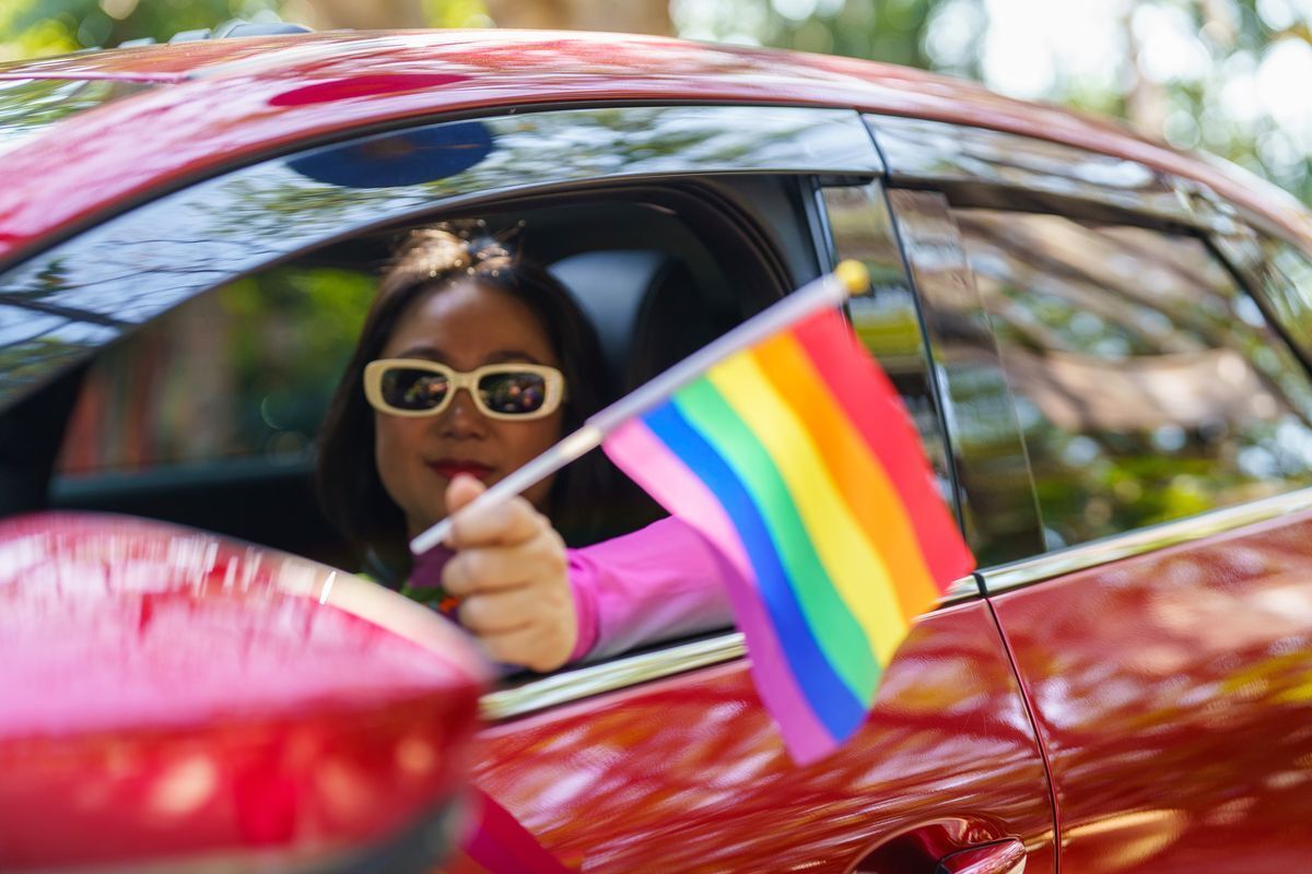 Asian woman in a car waving a Rainbow Flag.