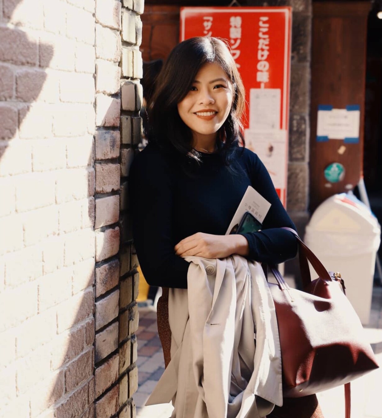 Asian woman happily holding a jacket, tickets, and a bag and leaning against a brick wall.