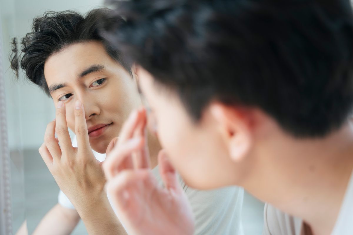 Asian man touching his nose in front of a mirror.