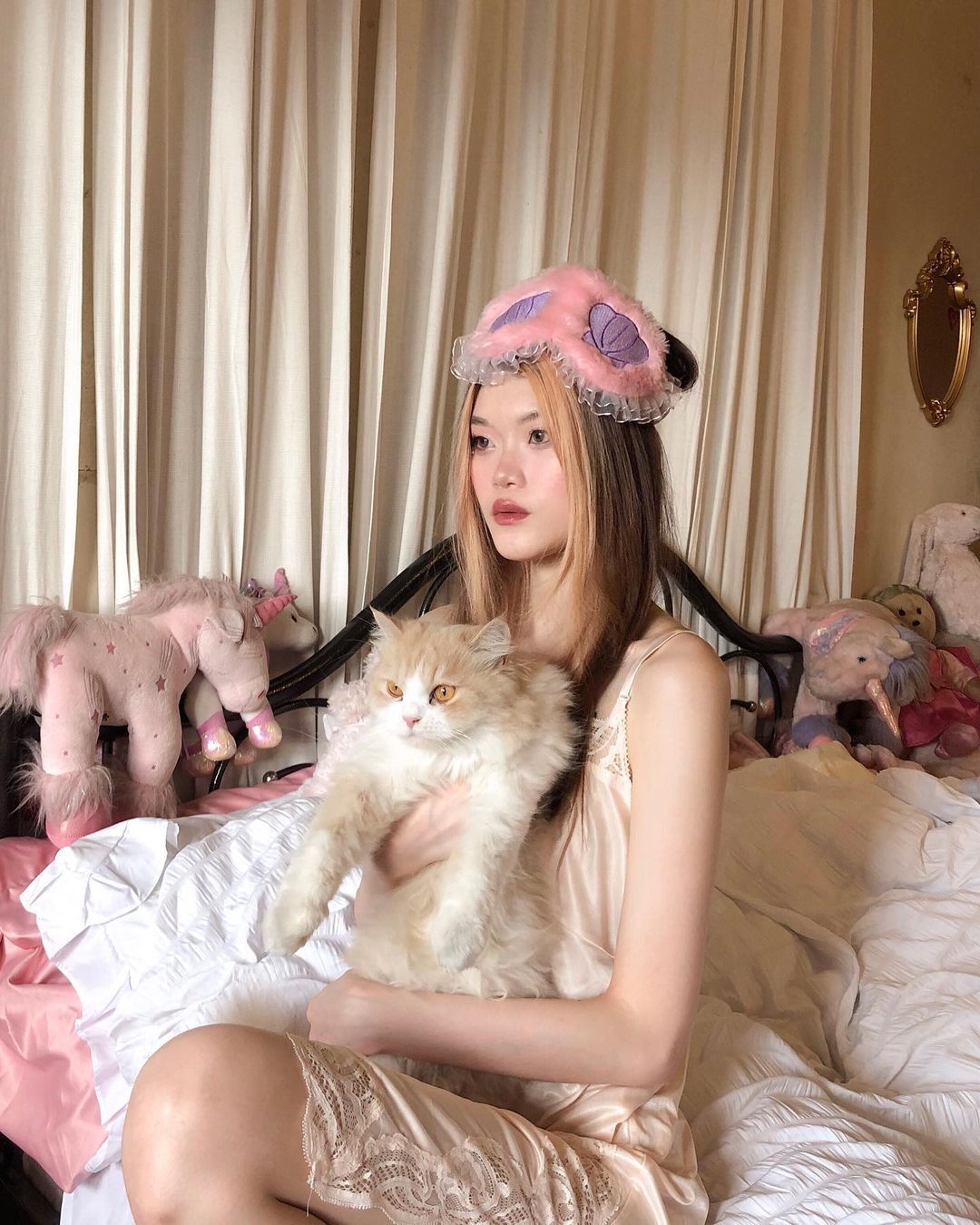 Woman with blonde moneypiece hair wearing silk nightdress and sleep mask while holding a cat.