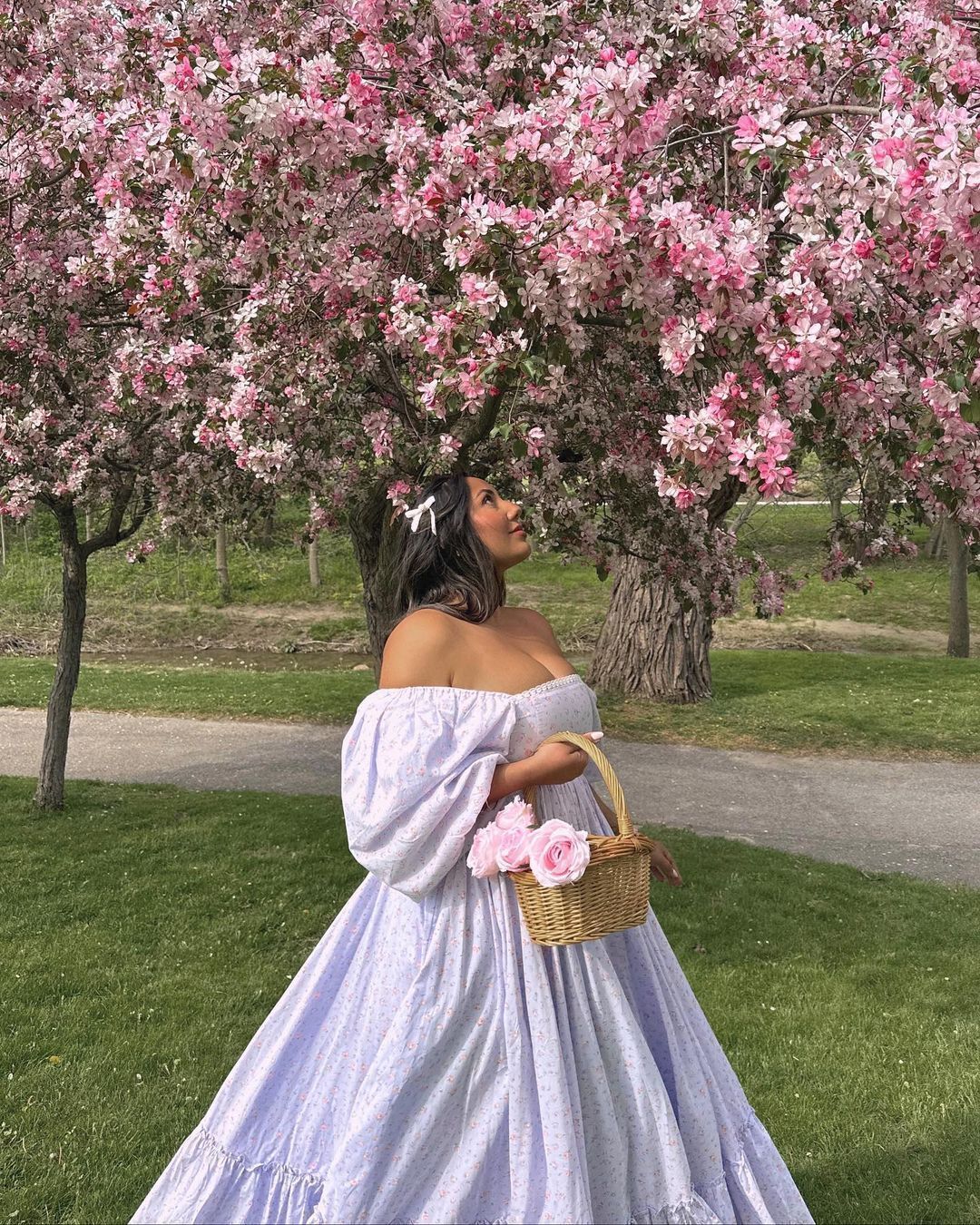 Woman wearing white off-shoulder dress while carrying hand basket filled with pink flowers
