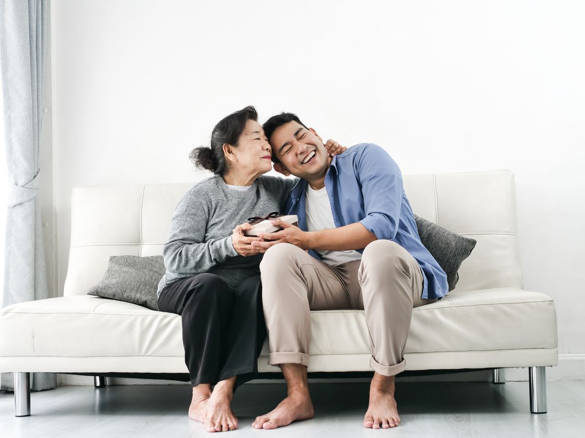 Old Asian woman happily putting her arm around a young smiling Asian man giving her a box while they’re both sitting on a white couch in a bright room.