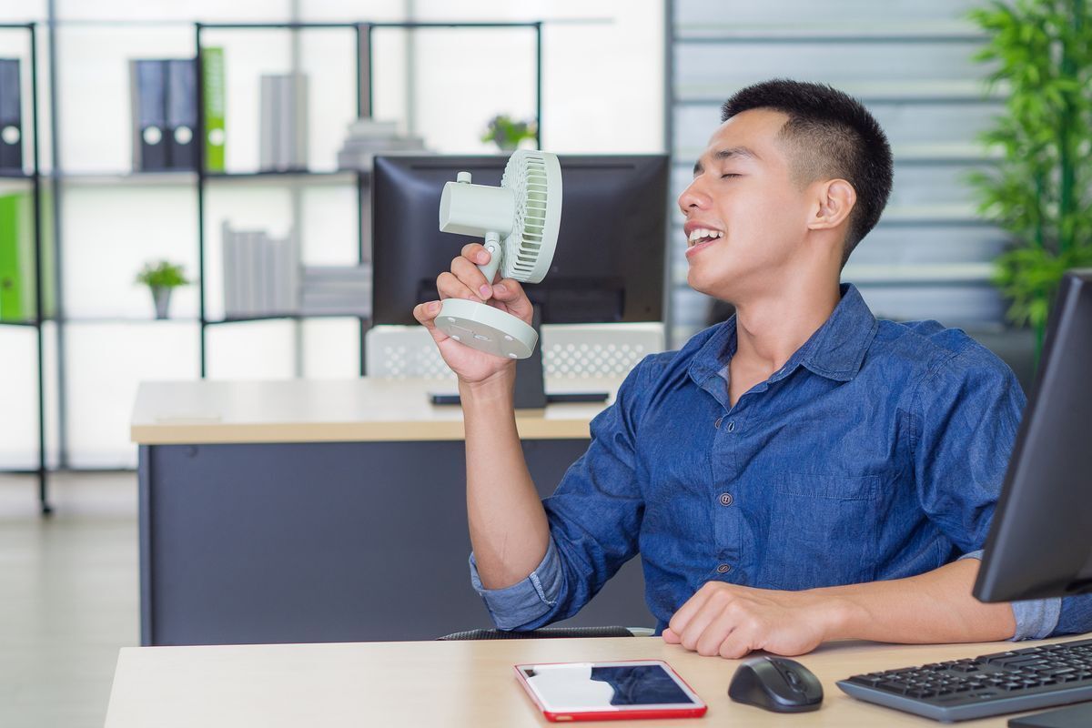 Man sitting behind his desk is comforting himself with a small portable electric fan.