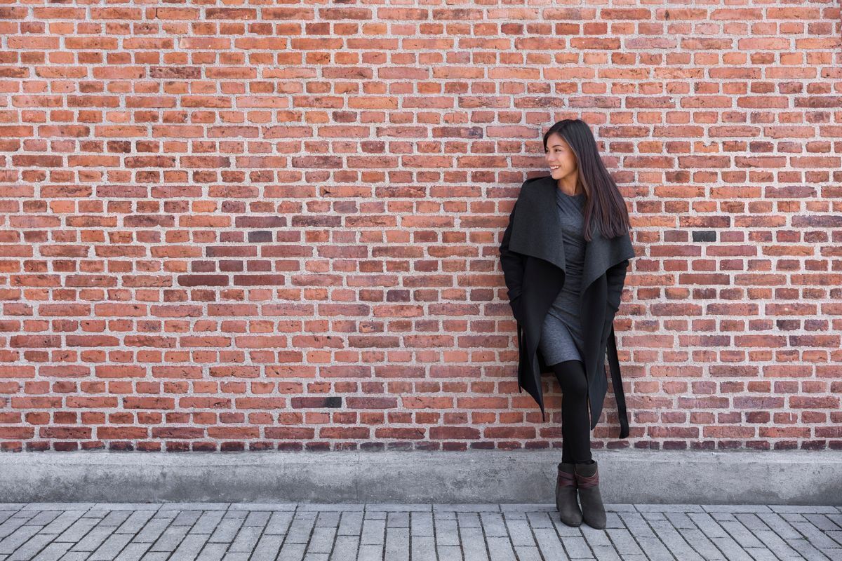 Asian woman in a black dress, tights, boots, and coat leaning on a brick wall