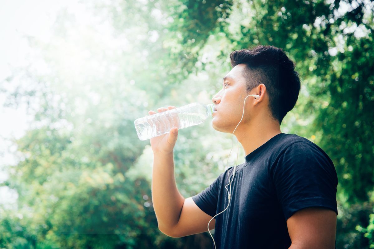 Asian man drinking from a bottle of water and wearing white earphones outdoors.