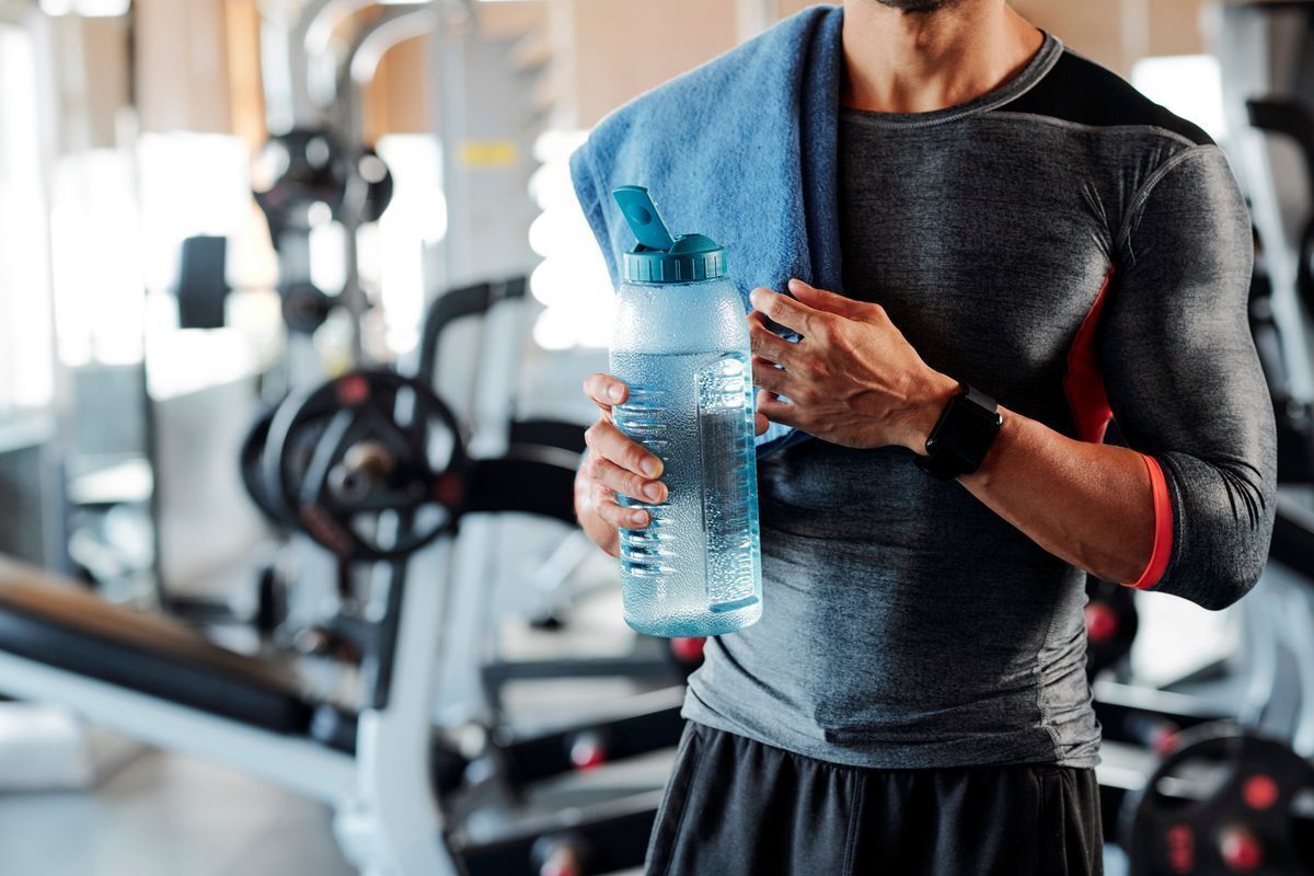 Cropped photo of a man’s torso wearing a rash guard with a towel draped over his shoulder and holding a water bottle.