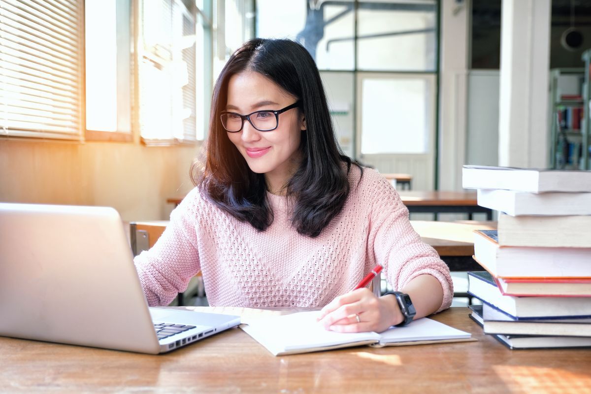 Asian woman in a pink sweater and glasses working on laptop and writing on a notebook with a stack of books on the table.