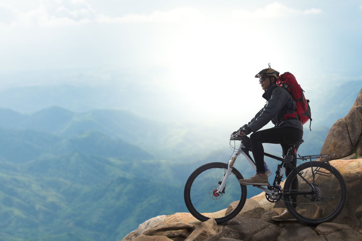 Man with a red backpack and bike on a mountain top looking out at a mountain.