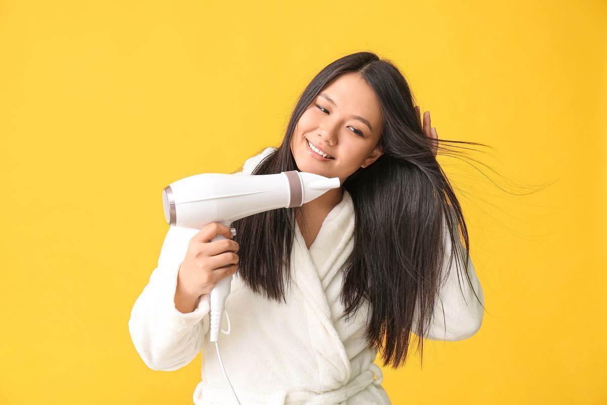 Asian woman in bathrobe blow drying her hair and smiling against a yellow background.