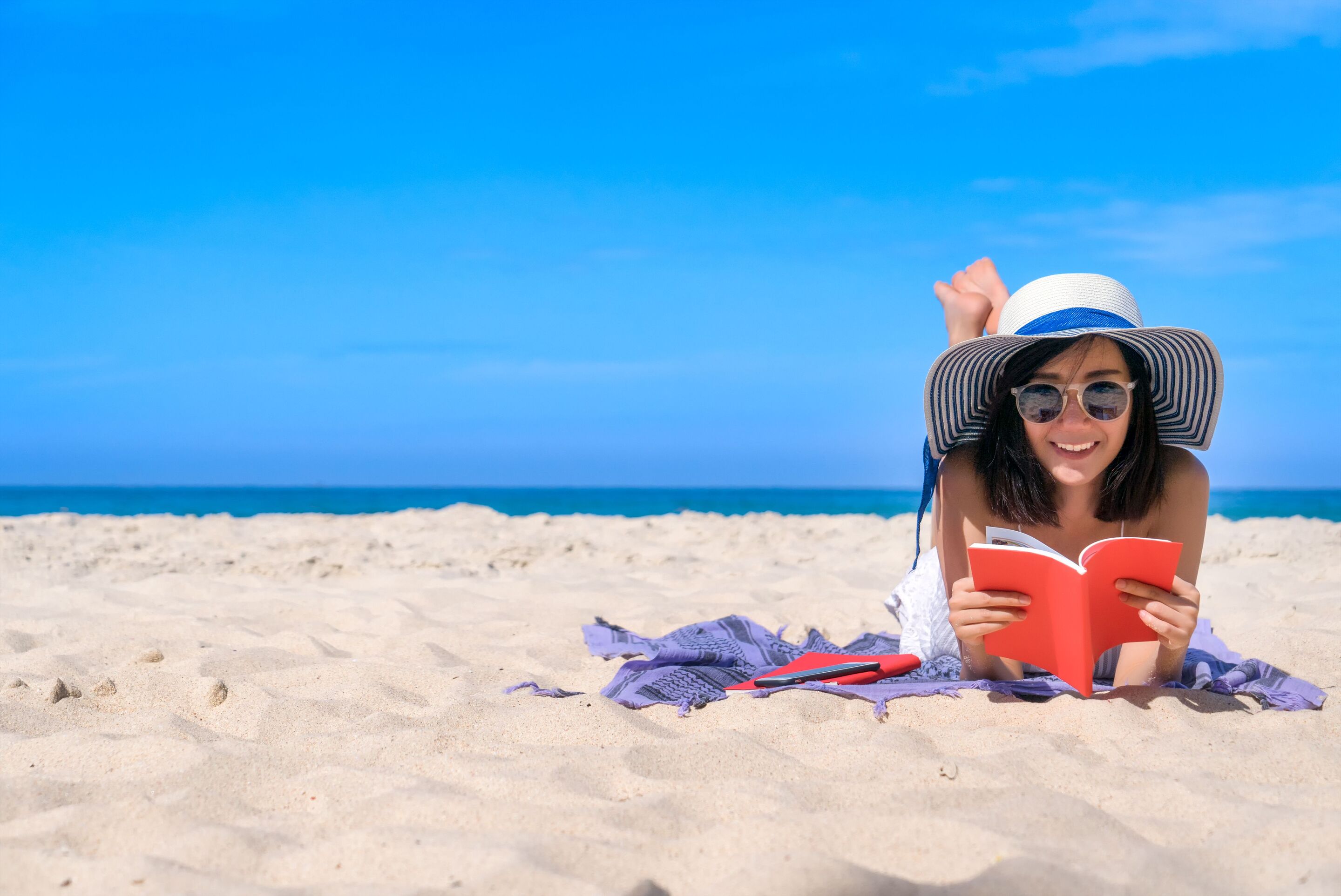 Asian woman wearing a sun hat smiling and reading a book on the beach