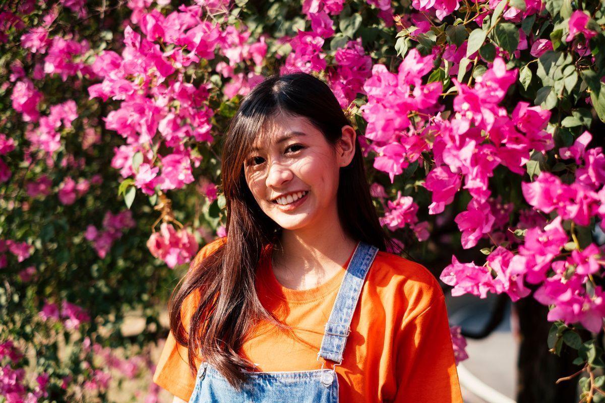 Young Asian woman in an orange shirt posing with flowers in the background.