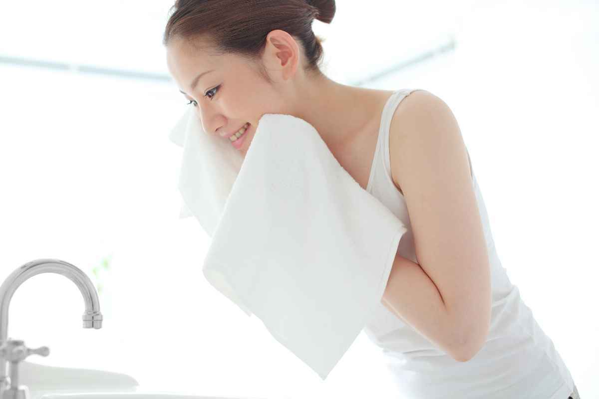 Woman patting her face with a white towel in front of a sink.